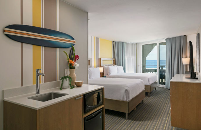 Beachy coastal hotel room with two queen beds, compact kitchenette with sink, microwave and mini-fridge, surfboard wall decor and fresh flowers, plus sliding glass doors opening to a balcony with ocean view.