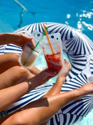 Two frozen cocktail pouches with colorful straws held over a navy-and-white inflatable pool float, tanned legs stretched out in sparkling blue pool water on a sunny summer day.