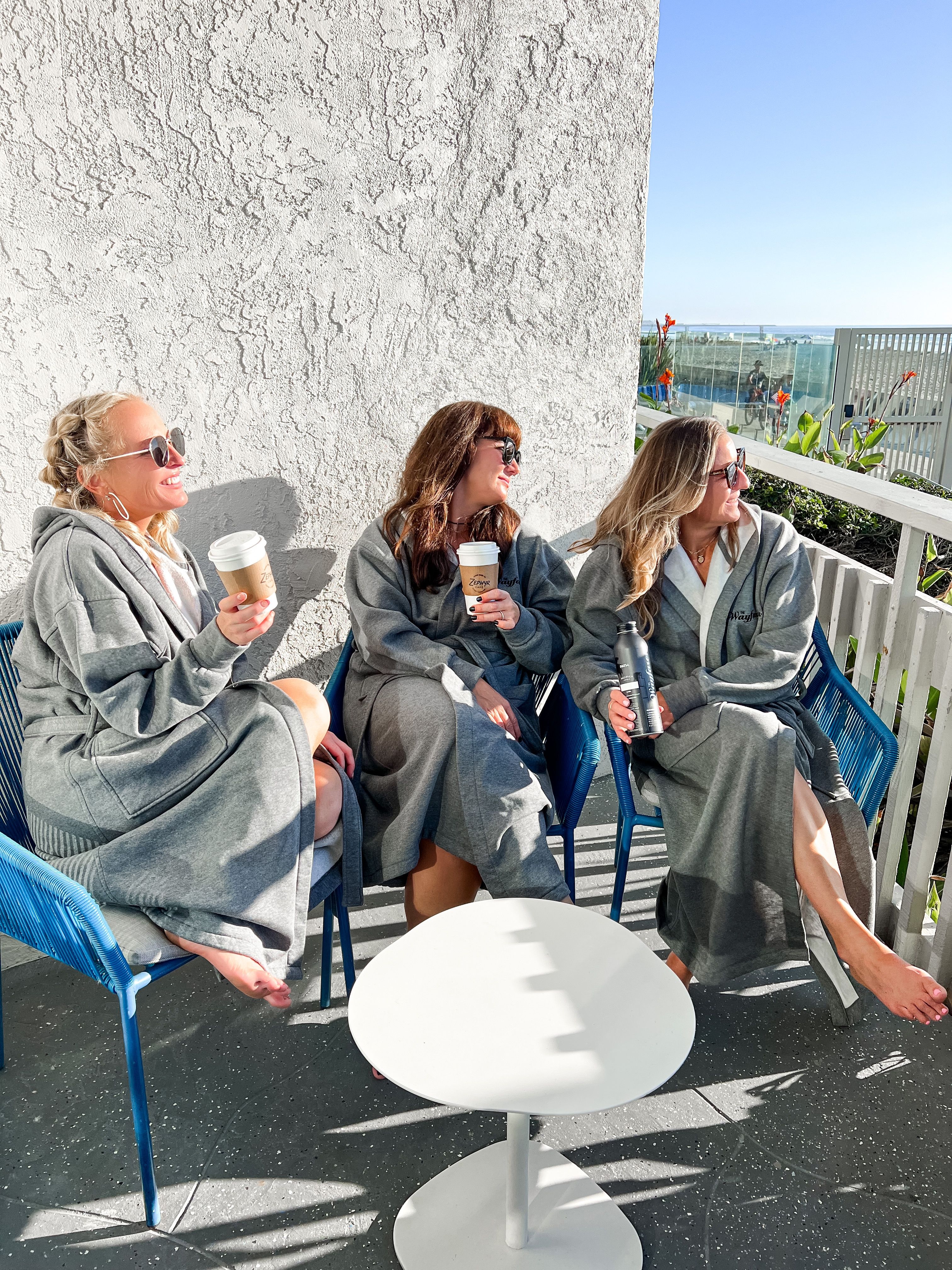 Three friends in matching gray robes relaxing barefoot on a sunny beachfront balcony, sipping coffee and a water bottle while wearing sunglasses with ocean-view and railing in the background