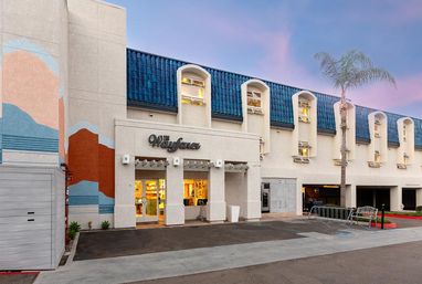Beachside boutique hotel exterior at dusk with blue tiled mansard roof and arched windows, colorful wall mural, lit glass entrance, palm tree, bike rack and parking area.