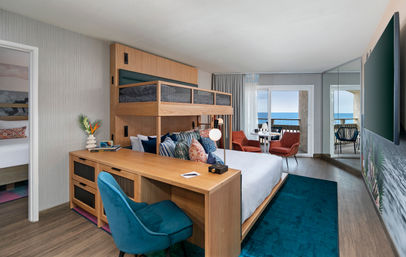 Beachfront hotel suite with ocean-view balcony, modern coastal decor, king bed with upper bunk, wood desk and teal chair, plus coral lounge chairs facing the sea.