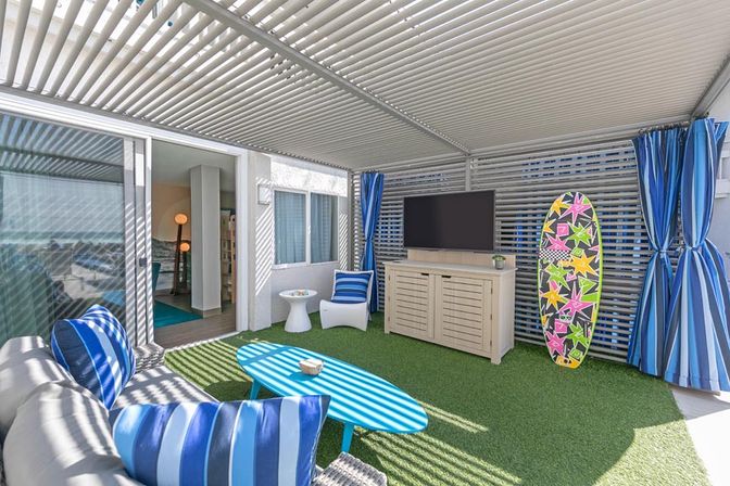 Covered coastal-style patio with slatted pergola casting striped shadows, blue-striped cushions on wicker sofa, turquoise coffee table, flat-screen TV, colorful surfboard decor, and artificial turf next to sliding glass door