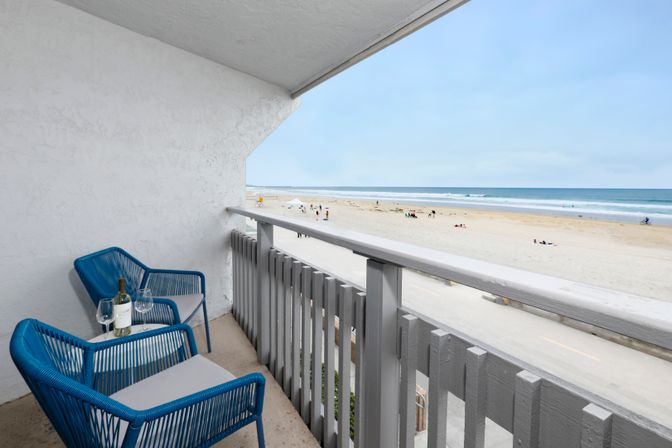 Oceanfront balcony with two blue wicker chairs and a bottle of wine on a small table, overlooking a sandy beach and gentle surf.