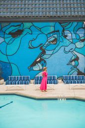 Woman in a bright pink dress and sunhat strolling poolside past striped blue lounge chairs and a turquoise pool beneath a large vibrant blue mural of abstract faces