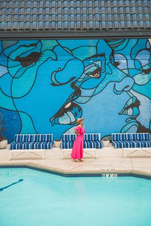 Woman in a bright pink dress and sunhat strolling poolside past striped blue lounge chairs and a turquoise pool beneath a large vibrant blue mural of abstract faces