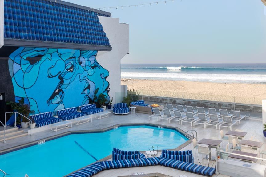 Oceanfront pool deck with blue-striped loungers and a large blue face mural on a building wall, overlooking sandy beach and rolling surf under a clear sky.