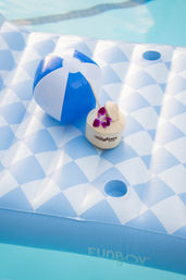 Blue-and-white inflatable pool float with a matching beach ball and a coconut drink garnished with a purple orchid, floating in a sunny swimming pool.