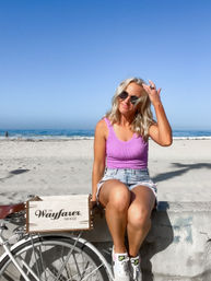 Woman in sunglasses and a pink tank top sitting on a seawall at a sunny San Diego beach, vintage-style bike with wooden crate nearby, ocean waves and clear blue sky behind her.