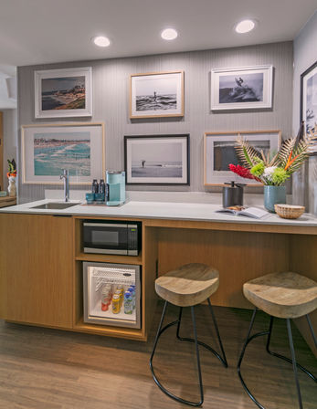 Coastal hotel kitchenette with framed beach photos on textured wall, white countertop with sink, microwave and stocked mini-fridge, wooden bar stools and colorful floral arrangement — modern seaside suite vibe.