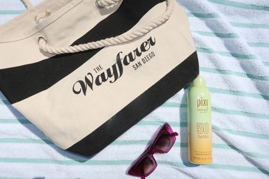 Beach-day essentials: black-and-cream striped canvas tote with rope handle, SPF 50 sun mist can, and fuchsia sunglasses on a teal-striped beach towel — San Diego coastal vibe