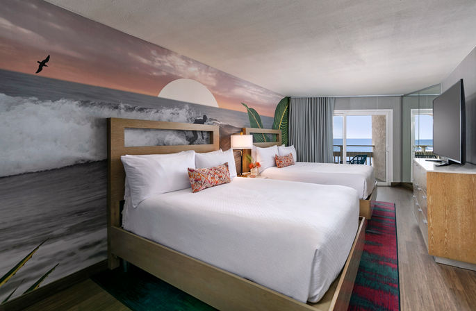 Ocean-view hotel room with two queen beds in white linens, colorful decorative pillows, coastal sunset mural on accent wall, balcony overlooking the sea, flat-screen TV and modern wood furniture.