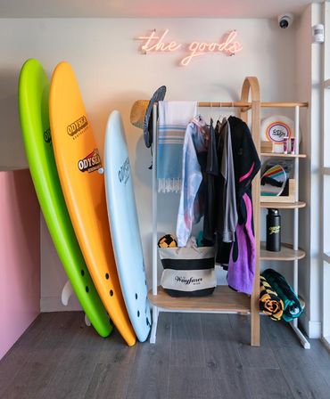 Colorful surf shop display: lime green, orange and light-blue surfboards leaning beside a wooden rack with beach towels, hats, boardshorts, a striped beach bag and rolled towels under a pink neon “the goods” sign.