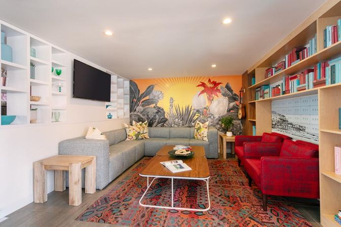 Bright modern living room with gray L-shaped sectional, red armchairs, colorful tropical sun mural, patterned rug, wooden coffee table and built-in shelves