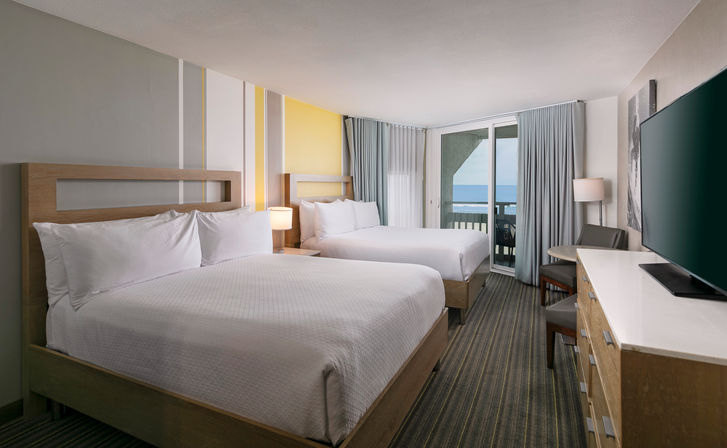 Coastal-modern ocean-view hotel room with two queen beds, light wood headboards, crisp white linens, bedside lamps, dresser with flat-screen TV and sliding balcony door overlooking the beach and sea.