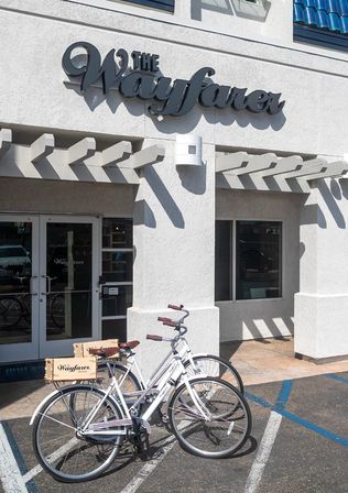 Sunlit white stucco boutique storefront with a cursive sign above a pergola casting striped shadows, and two vintage-style white cruiser bicycles with wooden cargo boxes parked by glass double doors.