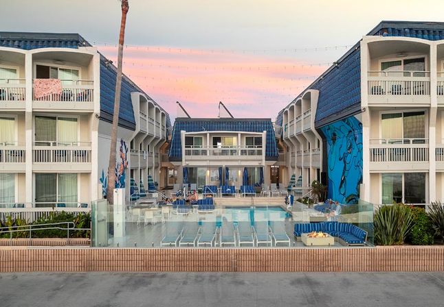 Beachside hotel courtyard with central swimming pool, blue-tiled roofs and balconies, palm tree, mural, string lights and poolside lounge chairs with a fire pit under a pink sunset sky.