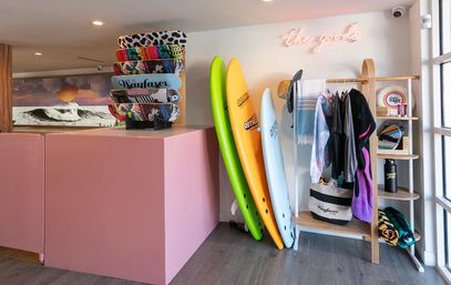 Vibrant coastal surf shop interior with a pastel pink counter, stacked skateboards, three colorful surfboards leaning on a rack, and beach apparel and accessories on shelves.