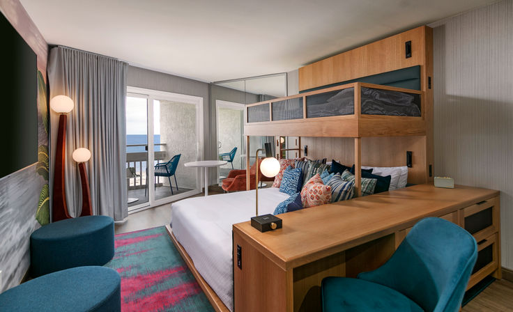 Modern ocean-view hotel room with king bed and built-in wooden bunk, colorful throw pillows, desk and seating, sliding doors to a balcony overlooking the beach
