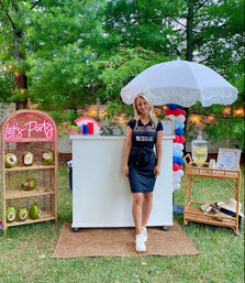Backyard summer party setup with a woman in an apron at a white pop-up bar under a fringed umbrella, red-white-blue balloon column, neon 'Let's Party' wicker shelf with coconuts, lemonade dispenser and rattan side table