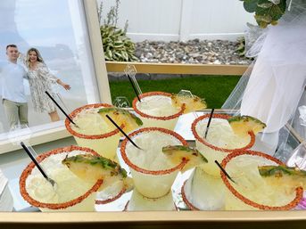 Mirrored tray of pineapple margaritas with chili-salt rims, pineapple wedge garnishes and black straws, with a framed portrait and white tulle in a backyard celebration setup.