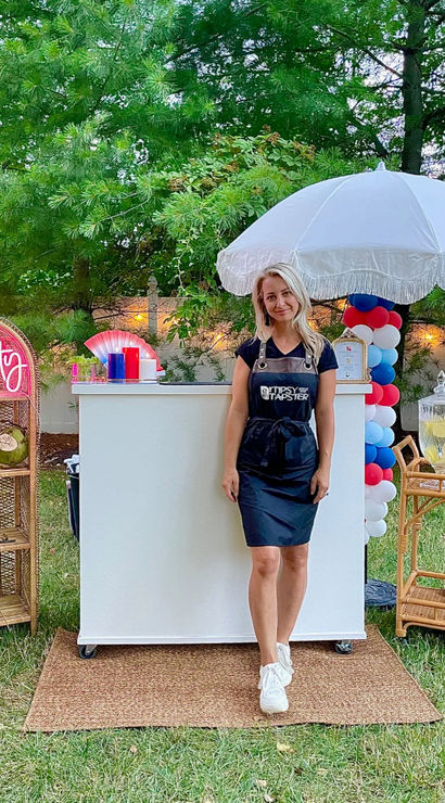Backyard summer party bar: woman in a black apron stands behind a white mobile bar under a fringed umbrella, red-white-blue balloon column, neon "Let's Party" sign, wicker shelves with coconuts and a lemonade dispenser on the grass.