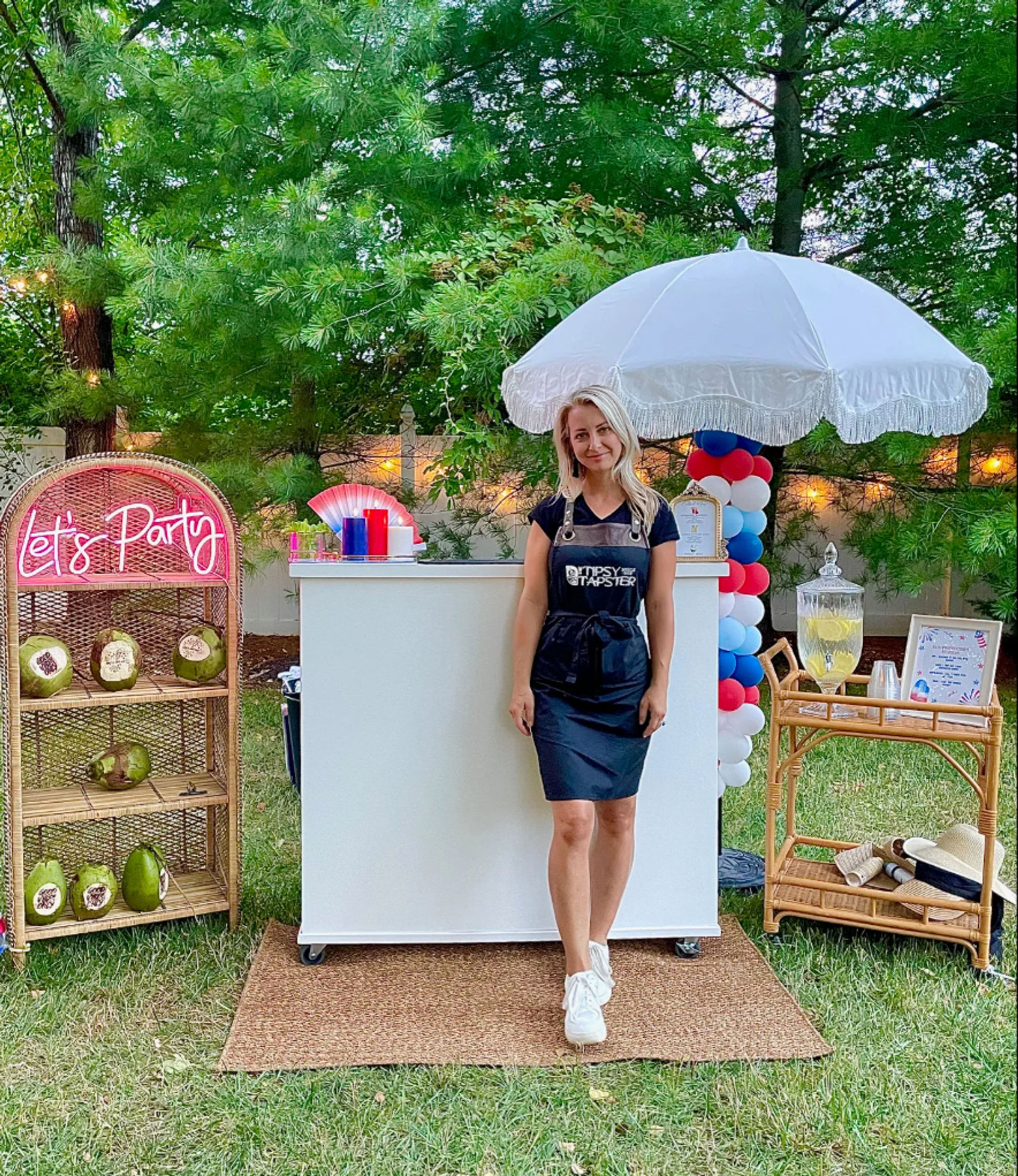Backyard summer party bar: woman in a black apron stands behind a white mobile bar under a fringed umbrella, red-white-blue balloon column, neon "Let's Party" sign, wicker shelves with coconuts and a lemonade dispenser on the grass.
