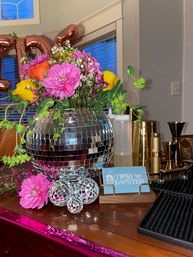 Vibrant floral centerpiece of pink and yellow blooms in a mirrored disco-ball vase with small mirrored orbs, sitting on an indoor party bar table surrounded by cocktail tools, gold jiggers and metallic balloons
