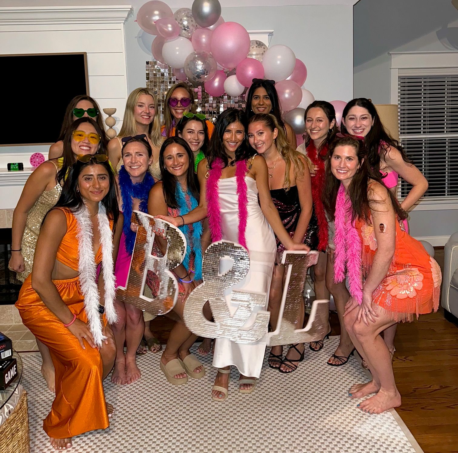Group of smiling women posing in a living room celebration with pink, white and silver balloon arch, colorful feather boas, and large glittery "B & L" letters.