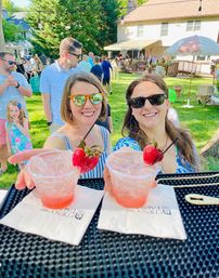 Two smiling women in sunglasses reaching for strawberry-garnished pink cocktails at a sunny backyard summer party, guests mingling on a grassy lawn and patio in the background.