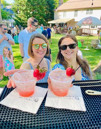 Two smiling women in sunglasses reaching for strawberry-garnished pink cocktails at a sunny backyard summer party, guests mingling on a grassy lawn and patio in the background.