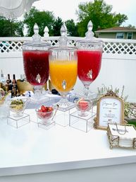 Backyard patio drink station with three ornate glass beverage dispensers filled with red and orange juices, small crystal bowls of fruit garnishes and a framed sign on a white table.