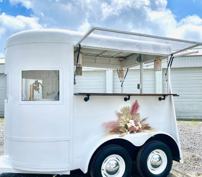 White vintage trailer converted into a mobile bar with an open serving window and wood counter, woven pendant lights hanging inside, pampas and dried floral arrangement on the wheel well, parked on gravel under a bright blue sky with fluffy clouds.