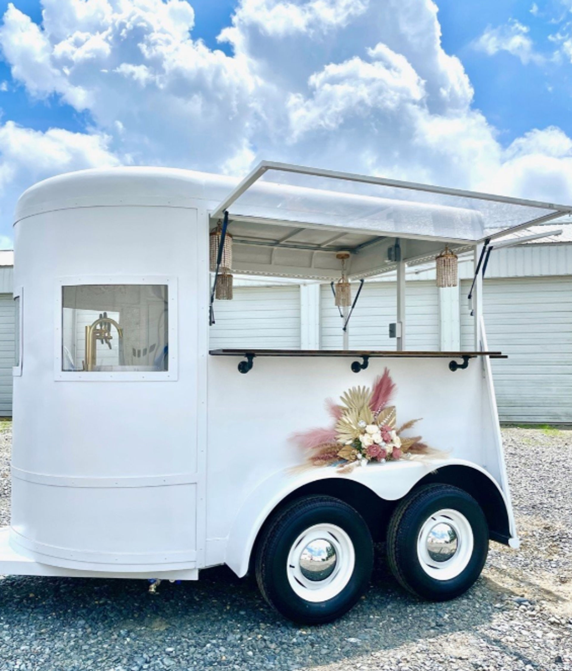 White vintage trailer converted into a mobile bar with an open serving window and wood counter, woven pendant lights hanging inside, pampas and dried floral arrangement on the wheel well, parked on gravel under a bright blue sky with fluffy clouds.
