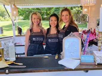Three smiling servers in black aprons behind a decorated outdoor mobile bar at a sunny garden event, with cocktail tools, a framed menu, colorful straws and glassware on the counter.