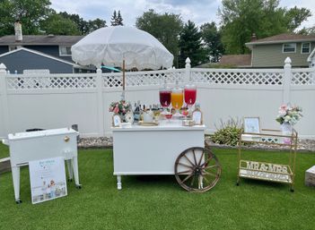 Chic backyard wedding drink station on green lawn: white umbrella-topped cart with three colorful beverage dispensers, floral arrangements, wooden wagon wheel accent, cooler and gold bar cart with “MR & MRS” sign in front of a white privacy fence.