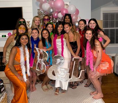 Group of smiling women in a living room at a festive indoor bachelorette-style party with pink and silver balloons, feather boas, and oversized sparkly B & L letters.