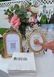Backyard party table with pink and white rose bouquet in a glass vase, ornate gold frames — one showing a QR social tag and the other a 'press for champagne' button being pressed, with champagne bottles and stacked napkins nearby.