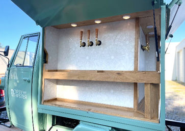 Mint-green vintage truck converted into a mobile bar, open side hatch revealing three brass beer taps above wooden shelving and a patterned white backsplash under recessed lights.