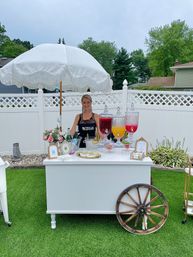 Backyard beverage cart under a white fringed umbrella on green lawn, attendant behind a white cart with three glass drink dispensers filled with red, orange and pink beverages, floral arrangement, glassware and a decorative wooden wagon wheel by a white lattice fence.