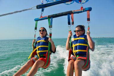 Two smiling women in blue life jackets tandem parasailing over turquoise coastal waters, gripping harness straps above a boat wake under a clear blue sky