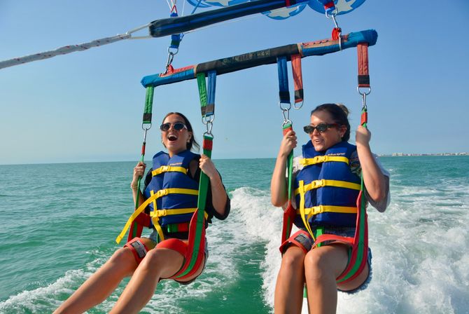 Two smiling women in blue life jackets tandem parasailing over turquoise coastal waters, gripping harness straps above a boat wake under a clear blue sky