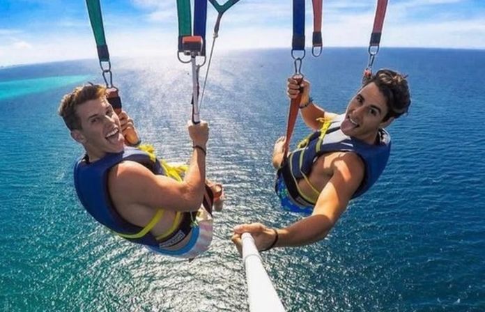 Two people parasailing in tandem over turquoise ocean, wearing life vests and smiling into a selfie stick with a sunny blue sky overhead