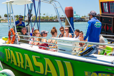 Group of passengers seated on a bright green parasailing boat in a sunny coastal harbor, crew briefing them at a waterfront pier with palm trees nearby.