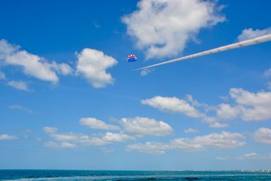Red-white-blue parasail soaring across a clear blue sky with puffy clouds above turquoise ocean and a distant shoreline.