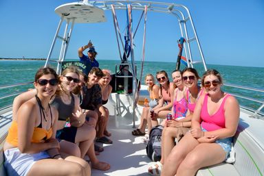 Smiling group of friends on a parasailing boat with a crew member flashing a shaka, turquoise ocean, clear blue sky, sunny coastal day trip
