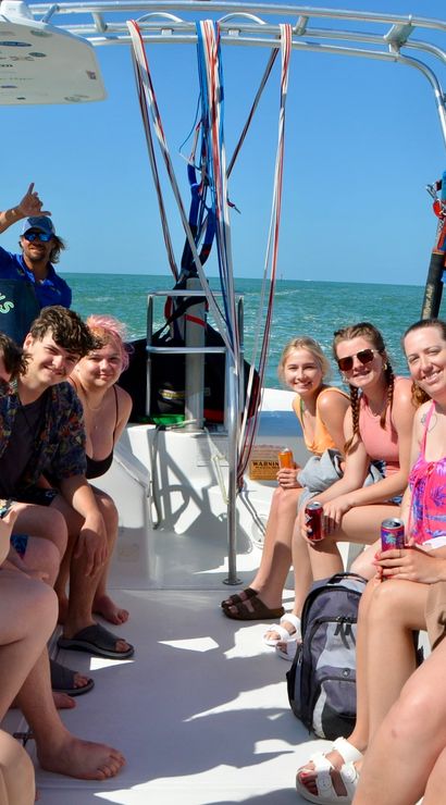 Smiling group of friends on a parasailing boat with a crew member flashing a shaka, turquoise ocean, clear blue sky, sunny coastal day trip