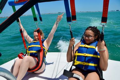 Two people in blue life jackets strapped into colorful parasailing harnesses on a boat, smiling and raising arms over turquoise ocean with a distant coastline and clear blue sky.