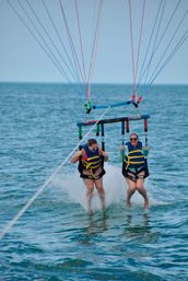 Two people tandem parasailing over turquoise ocean, wearing yellow life jackets and sunglasses with feet skimming the water and splashes beneath colorful parachute lines.