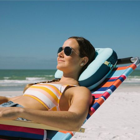 Person in striped bikini top wearing aviator sunglasses, reclining on a colorful beach chair with a neck pillow, sunny ocean beach in background