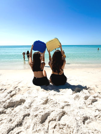 Two people sitting on white-sand beach facing turquoise ocean under a clear blue sky, holding bright blue and yellow beach bags overhead for a sunny summer day.